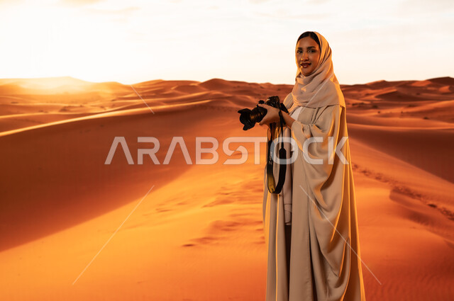 Artistic photography shots, practicing a favorite hobby with passion, a professional photographer taking souvenir photos using modern techniques, soft golden sand, a young Saudi Arabian Gulf woman wearing a colorful abaya standing in the desert areas holding a high-resolution camera in her hands