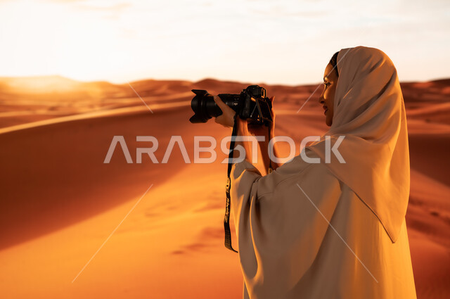 Soft golden sand, artistic photography shots, practicing a favorite hobby with passion, a professional photographer taking souvenir photos using modern techniques, a close-up from the back of a young Saudi Arabian Gulf woman wearing a colorful abaya standing in the desert areas holding a high-resolution camera in her hands