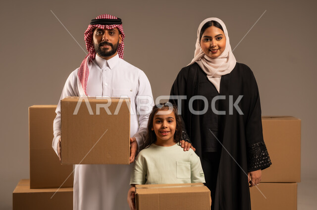 صناديق وحاويات كرتونية مليئة بالأغراض المنزلية، مفهوم الانتقال إلى سكن جديد، بورتريه مقرب لعائلة عربية سعودية خليجية تقف باستقامة بتعابير السرور، الاعتناء بالأطفال والاهتمام بهم, قضاء أوقات ممتعة مع الاسرة، خلفيه رمادية