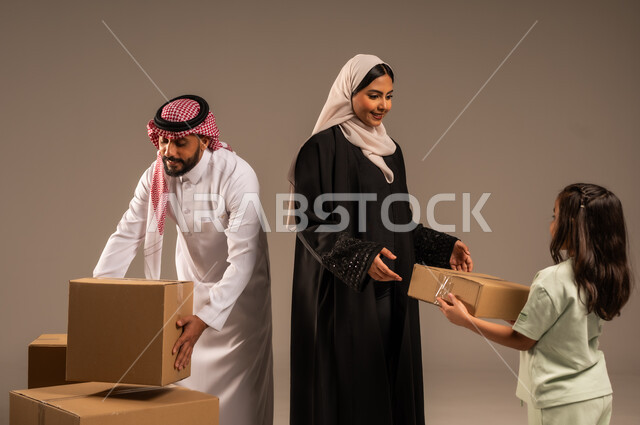 صناديق وحاويات كرتونية مليئة بالأغراض المنزلية، مفهوم الانتقال إلى سكن جديد، بورتريه مقرب لعائلة عربية سعودية خليجية تقف باستقامة بتعابير السرور، الاعتناء بالأطفال والاهتمام بهم, قضاء أوقات ممتعة مع الاسرة، خلفيه رمادية