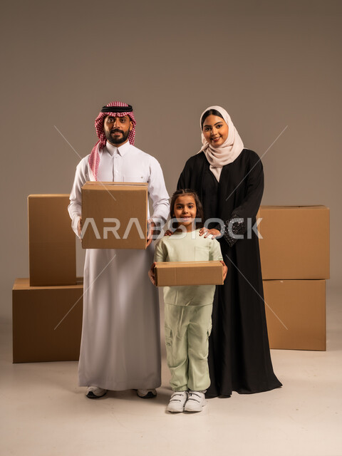 مفهوم الانتقال إلى سكن جديد، صناديق وحاويات كرتونية مليئة بالأغراض المنزلية، بورتريه لعائلة عربية سعودية خليجية تقف باستقامة بتعابير السرور، الاعتناء بالأطفال والاهتمام بهم, قضاء أوقات ممتعة مع الاسرة، الجسم بالطول الكامل، خلفيه رمادية