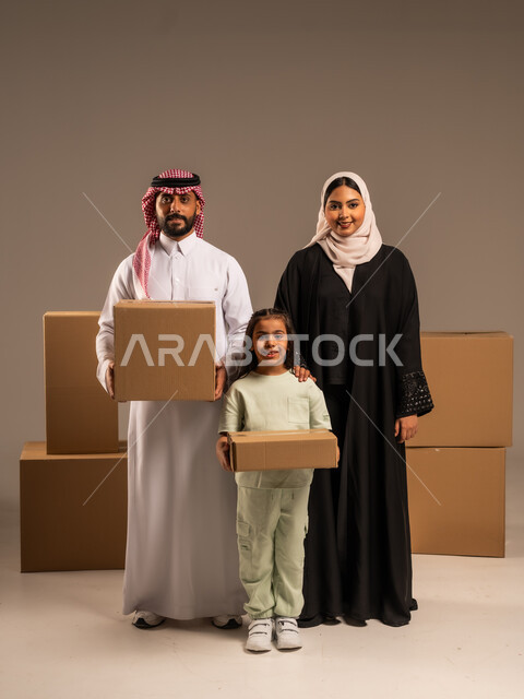 مفهوم الانتقال إلى سكن جديد، صناديق وحاويات كرتونية مليئة بالأغراض المنزلية، بورتريه لعائلة عربية سعودية خليجية تقف باستقامة بتعابير السرور، الاعتناء بالأطفال والاهتمام بهم, قضاء أوقات ممتعة مع الاسرة، الجسم بالطول الكامل، خلفيه رمادية