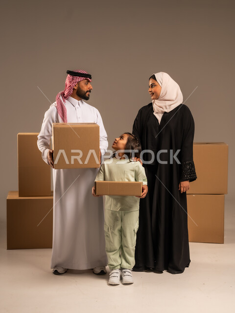 مفهوم الانتقال إلى سكن جديد، صناديق وحاويات كرتونية مليئة بالأغراض المنزلية، بورتريه لعائلة عربية سعودية خليجية تقف باستقامة بتعابير السرور، الاعتناء بالأطفال والاهتمام بهم, قضاء أوقات ممتعة مع الاسرة، الجسم بالطول الكامل، خلفيه رمادية