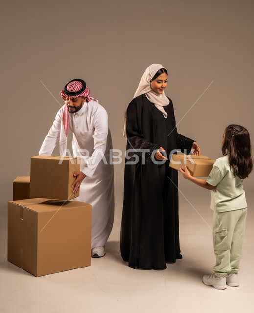 مفهوم الانتقال إلى سكن جديد، صناديق وحاويات كرتونية مليئة بالأغراض المنزلية، بورتريه لعائلة عربية سعودية خليجية تقف باستقامة بتعابير السرور، الاعتناء بالأطفال والاهتمام بهم, قضاء أوقات ممتعة مع الاسرة، الجسم بالطول الكامل، خلفيه رمادية
