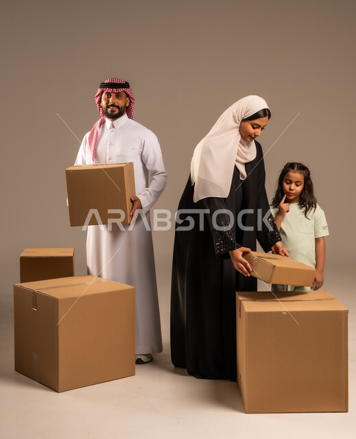 مفهوم الانتقال إلى سكن جديد، صناديق وحاويات كرتونية مليئة بالأغراض المنزلية، بورتريه لعائلة عربية سعودية خليجية تقف باستقامة بتعابير السرور، الاعتناء بالأطفال والاهتمام بهم, قضاء أوقات ممتعة مع الاسرة، الجسم بالطول الكامل، خلفيه رمادية