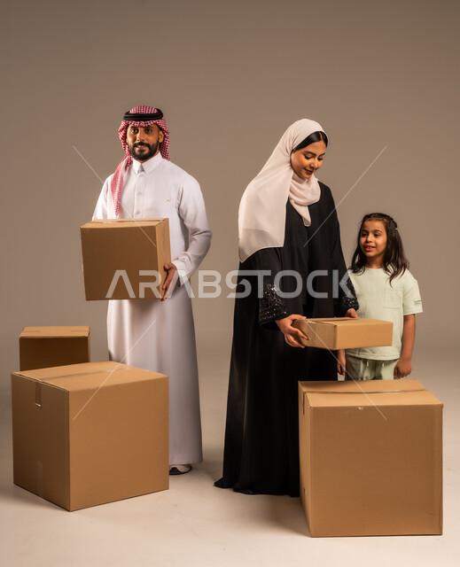 مفهوم الانتقال إلى سكن جديد، صناديق وحاويات كرتونية مليئة بالأغراض المنزلية، بورتريه لعائلة عربية سعودية خليجية تقف باستقامة بتعابير السرور، الاعتناء بالأطفال والاهتمام بهم, قضاء أوقات ممتعة مع الاسرة، الجسم بالطول الكامل، خلفيه رمادية