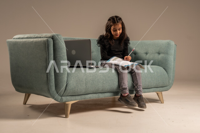 تلقي المحاضرات المدرسيه عبر الانترنت، متابعة الدروس أونلاين، مفهوم التعلم عن بعد باستخدام الأجهزة التقنية الحديثة والتكنولوجيا، بورتريه لفتاة عربية خليجية سعودية ترتدي الزي الكاجوال تجلس على كرسي مريح أمام الحاسوب المحمول,  خلفية رمادية