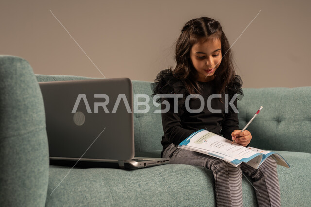 تلقي المحاضرات المدرسيه عبر الانترنت، متابعة الدروس أونلاين، مفهوم التعلم عن بعد باستخدام الأجهزة التقنية الحديثة والتكنولوجيا، بورتريه لفتاة عربية خليجية سعودية ترتدي الزي الكاجوال تجلس على كرسي مريح أمام الحاسوب المحمول,  خلفية رمادية