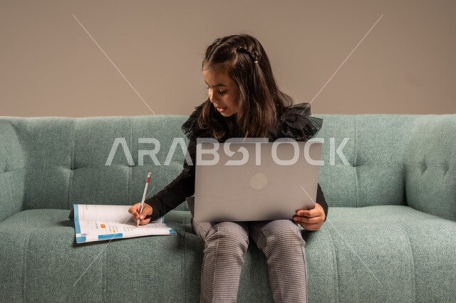تلقي المحاضرات المدرسيه عبر الانترنت، متابعة الدروس أونلاين، مفهوم التعلم عن بعد باستخدام الأجهزة التقنية الحديثة والتكنولوجيا، بورتريه لفتاة عربية خليجية سعودية ترتدي الزي الكاجوال تجلس على كرسي مريح أمام الحاسوب المحمول,  خلفية رمادية
