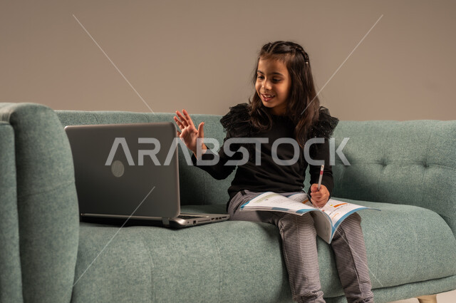  متابعة الدروس أونلاين، مفهوم التعلم عن بعد باستخدام الأجهزة التقنية الحديثة والتكنولوجيا، بورتريه لفتاة عربية خليجية سعودية ترتدي الزي الكاجوال تجلس على كرسي مريح أمام الحاسوب المحمول ترفع يدها للأعلى بايماءات القاء التحية والسلام,  خلفية رمادية