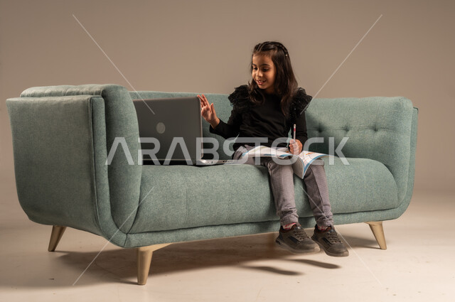 Follow lessons online, the concept of distance learning using modern technical devices and technology, a portrait of a Saudi Arabian Gulf girl wearing casual clothes sitting on a comfortable chair in front of a laptop raising her hand up with gestures of greeting and peace, gray background