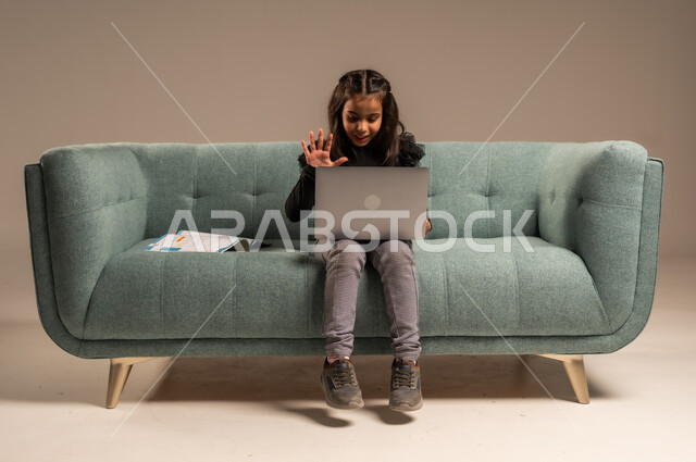  متابعة الدروس أونلاين، مفهوم التعلم عن بعد باستخدام الأجهزة التقنية الحديثة والتكنولوجيا، بورتريه لفتاة عربية خليجية سعودية ترتدي الزي الكاجوال تجلس على كرسي مريح أمام الحاسوب المحمول ترفع يدها للأعلى بايماءات القاء التحية والسلام,  خلفية رمادية