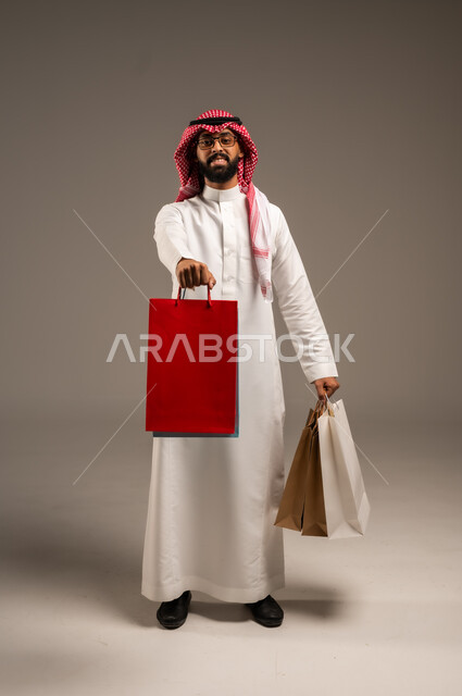 التبضع وأخذ الحاجات اللازمة, المشتريات التجارية والهدايا, مواسم التخفيضات في السعودية, الاستفادة من الحسومات والتصفيات, بورتريه لشاب عربي خليجي سعودي يرتدي الثوب التقليدي والشماغ يمسك بيده أكياس ورقية ملونة, متعة التسوق في المملكة, الجسم بالطول الكامل, خلفية رمادية