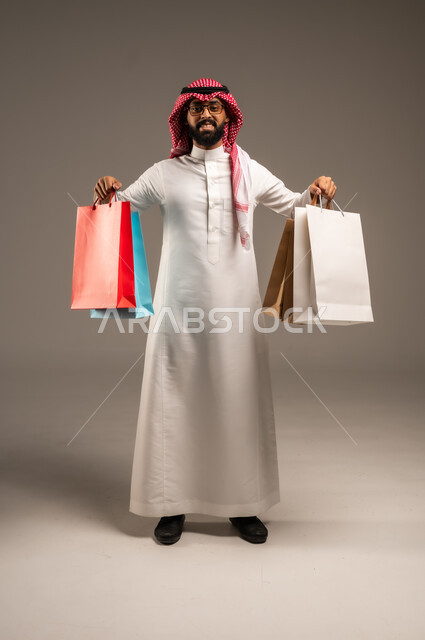التبضع وأخذ الحاجات اللازمة, المشتريات التجارية والهدايا, مواسم التخفيضات في السعودية, الاستفادة من الحسومات والتصفيات, بورتريه لشاب عربي خليجي سعودي يرتدي الثوب التقليدي والشماغ يمسك بيده أكياس ورقية ملونة, متعة التسوق في المملكة, الجسم بالطول الكامل, خلفية رمادية