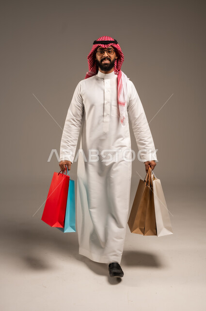 التبضع وأخذ الحاجات اللازمة, المشتريات التجارية والهدايا, مواسم التخفيضات في السعودية, الاستفادة من الحسومات والتصفيات, بورتريه لشاب عربي خليجي سعودي يرتدي الثوب التقليدي والشماغ يمسك بيده أكياس ورقية ملونة, متعة التسوق في المملكة, الجسم بالطول الكامل, خلفية رمادية