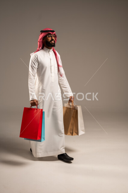 Shopping and taking the necessary needs, commercial purchases and gifts, discount seasons in Saudi Arabia, taking advantage of discounts and liquidations, portrait of a young Saudi Arabian Gulf man wearing the traditional dress and shemagh holding colorful paper bags in his hand, the joy of shopping in the Kingdom, full-length body, gray background