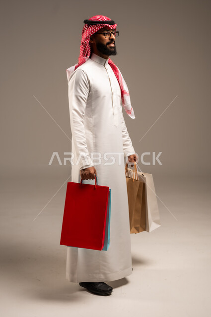 Shopping and taking the necessary needs, commercial purchases and gifts, discount seasons in Saudi Arabia, taking advantage of discounts and liquidations, portrait of a young Saudi Arabian Gulf man wearing the traditional dress and shemagh holding colorful paper bags in his hand, the joy of shopping in the Kingdom, full-length body, gray background
