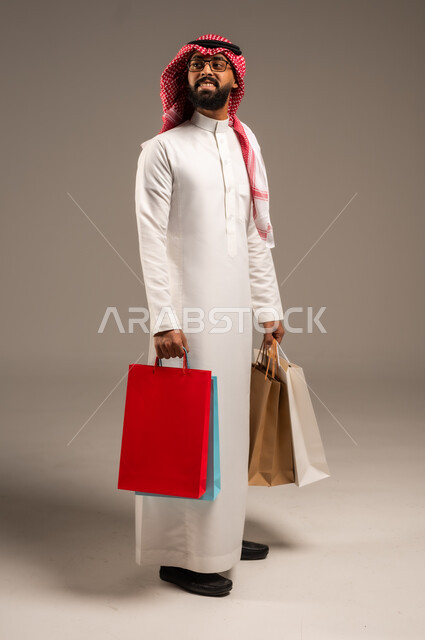 Shopping and taking the necessary needs, commercial purchases and gifts, discount seasons in Saudi Arabia, taking advantage of discounts and liquidations, portrait of a young Saudi Arabian Gulf man wearing the traditional dress and shemagh holding colorful paper bags in his hand, the joy of shopping in the Kingdom, full-length body, gray background