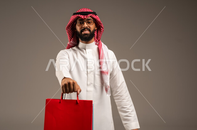 The joy of shopping in the Kingdom, shopping and taking the necessary needs, commercial purchases and gifts, discount seasons in Saudi Arabia, taking advantage of discounts and liquidations, close-up portrait of a young Saudi Arabian Gulf man wearing the traditional dress and shemagh holding colorful paper bags in his hand, gray background