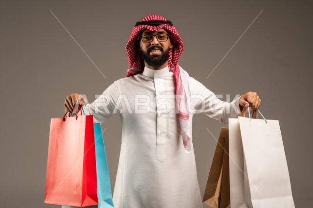 متعة التسوق في المملكة, التبضع وأخذ الحاجات اللازمة, المشتريات التجارية والهدايا, مواسم التخفيضات في السعودية, الاستفادة من الحسومات والتصفيات, بورتريه مقرب لشاب عربي خليجي سعودي يرتدي الثوب التقليدي والشماغ يمسك بيده أكياس ورقية ملونة, خلفية رمادية