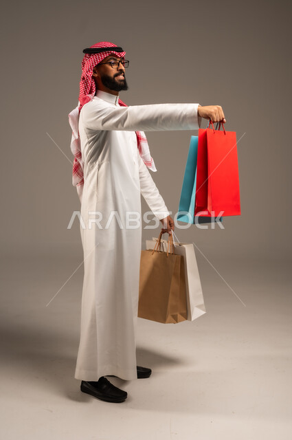 مواسم التخفيضات في السعودية, الاستفادة من الحسومات والتصفيات, المشتريات التجارية والهدايا, متعة التسوق في المملكة, التبضع وأخذ الحاجات اللازمة, بورتريه من الجانب لشاب عربي خليجي سعودي يرتدي الثوب التقليدي والشماغ يمسك بيده أكياس ورقية ملونة, خلفية رمادية