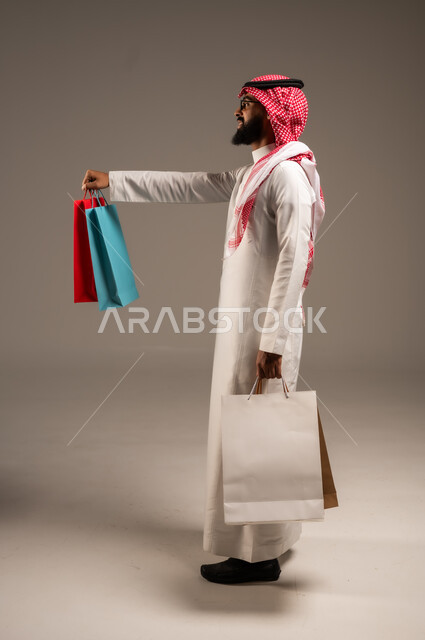 Discount seasons in Saudi Arabia, taking advantage of discounts and liquidations, commercial purchases and gifts, the pleasure of shopping in the Kingdom, shopping and taking the necessary necessities, a side portrait of a young Saudi Arabian Gulf man wearing the traditional dress and shemagh holding colored paper bags in his hand, gray background