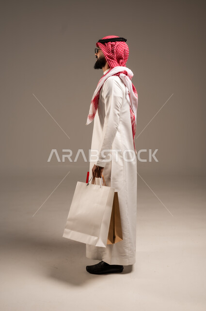 Discount seasons in Saudi Arabia, taking advantage of discounts and liquidations, commercial purchases and gifts, the pleasure of shopping in the Kingdom, shopping and taking the necessary necessities, a side portrait of a young Saudi Arabian Gulf man wearing the traditional dress and shemagh holding colored paper bags in his hand, gray background