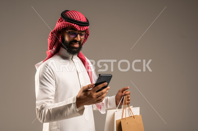Completing the order process using a mobile phone, the concept of remote shopping, online electronic purchases, close-up portrait of a young Saudi Arabian Gulf man wearing a traditional thobe and shemagh holding colorful paper bags in his hand, shopping and taking advantage of offers and discounts, gray background