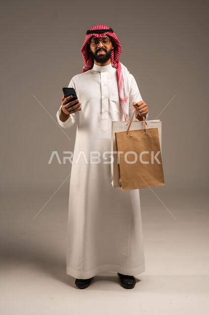 عمليات الشراء الالكتروني عبر الانترنت, إتمام عملية الطلبات باستخدام الهاتف المحمول، مفهوم التسوق عن بعد, بورتريه لشاب عربي خليجي سعودي يرتدي الثوب التقليدي والشماغ يمسك بيده أكياس ورقية ملونة, التبضع والاستفادة من العروض والتخفيضات, الجسم بالطول الكامل، خلفية رمادية