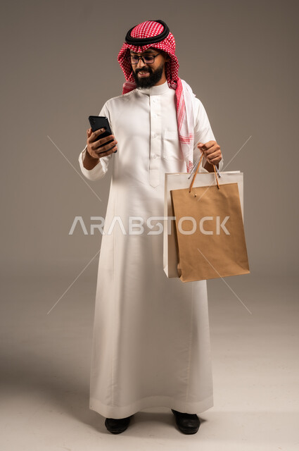 عمليات الشراء الالكتروني عبر الانترنت, إتمام عملية الطلبات باستخدام الهاتف المحمول، مفهوم التسوق عن بعد, بورتريه لشاب عربي خليجي سعودي يرتدي الثوب التقليدي والشماغ يمسك بيده أكياس ورقية ملونة, التبضع والاستفادة من العروض والتخفيضات, الجسم بالطول الكامل، خلفية رمادية