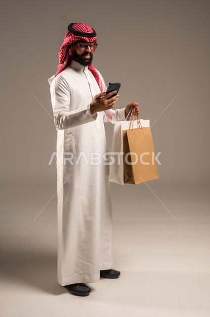 عمليات الشراء الالكتروني عبر الانترنت, إتمام عملية الطلبات باستخدام الهاتف المحمول، مفهوم التسوق عن بعد, بورتريه لشاب عربي خليجي سعودي يرتدي الثوب التقليدي والشماغ يمسك بيده أكياس ورقية ملونة, التبضع والاستفادة من العروض والتخفيضات, الجسم بالطول الكامل، خلفية رمادية