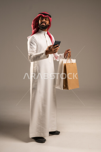 عمليات الشراء الالكتروني عبر الانترنت, إتمام عملية الطلبات باستخدام الهاتف المحمول، مفهوم التسوق عن بعد, بورتريه لشاب عربي خليجي سعودي يرتدي الثوب التقليدي والشماغ يمسك بيده أكياس ورقية ملونة, التبضع والاستفادة من العروض والتخفيضات, الجسم بالطول الكامل، خلفية رمادية