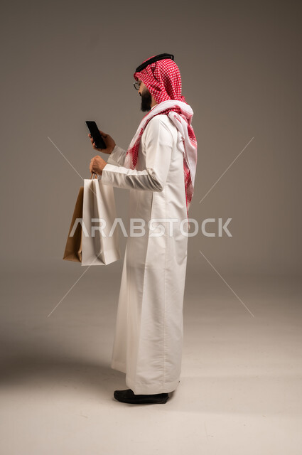 عمليات الشراء الالكتروني عبر الانترنت, إتمام عملية الطلبات باستخدام الهاتف المحمول، مفهوم التسوق عن بعد, بورتريه لشاب عربي خليجي سعودي يرتدي الثوب التقليدي والشماغ يمسك بيده أكياس ورقية ملونة, التبضع والاستفادة من العروض والتخفيضات, الجسم بالطول الكامل، خلفية رمادية