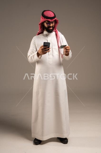 إمكانية تسديد الفواتير بالبطاقات الإلكترونية, استخدام الأجهزة والتقنيات الحديثة, سهولة الشراء والدفع عن طريق بطاقة الإئتمان البنكية، بورتريه لرجل عربي خليجي سعودي مبتسم يرتدي الثوب التقليدي والشماغ يمسك بيده بطاقة الصراف ويعمل على الهاتف المحمول, خلفية رمادية