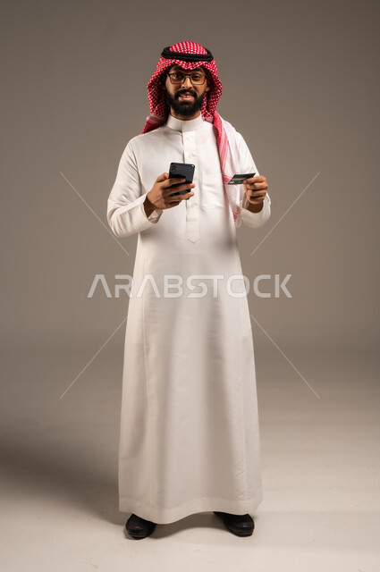 إمكانية تسديد الفواتير بالبطاقات الإلكترونية, استخدام الأجهزة والتقنيات الحديثة, سهولة الشراء والدفع عن طريق بطاقة الإئتمان البنكية، بورتريه لرجل عربي خليجي سعودي مبتسم يرتدي الثوب التقليدي والشماغ يمسك بيده بطاقة الصراف ويعمل على الهاتف المحمول, خلفية رمادية