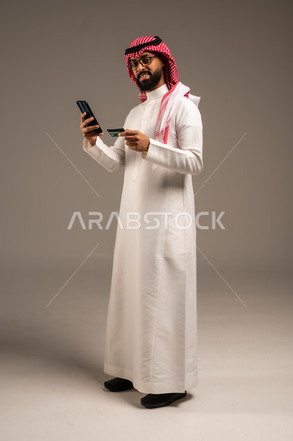 إمكانية تسديد الفواتير بالبطاقات الإلكترونية, استخدام الأجهزة والتقنيات الحديثة, سهولة الشراء والدفع عن طريق بطاقة الإئتمان البنكية، بورتريه لرجل عربي خليجي سعودي مبتسم يرتدي الثوب التقليدي والشماغ يمسك بيده بطاقة الصراف ويعمل على الهاتف المحمول, خلفية رمادية