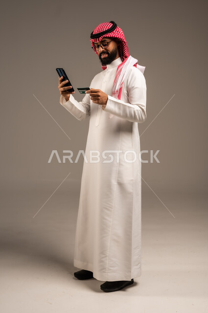 إمكانية تسديد الفواتير بالبطاقات الإلكترونية, استخدام الأجهزة والتقنيات الحديثة, سهولة الشراء والدفع عن طريق بطاقة الإئتمان البنكية، بورتريه لرجل عربي خليجي سعودي مبتسم يرتدي الثوب التقليدي والشماغ يمسك بيده بطاقة الصراف ويعمل على الهاتف المحمول, خلفية رمادية