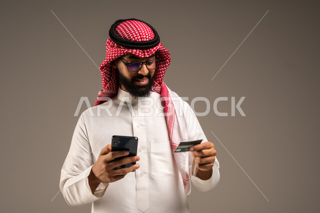  استخدام الأجهزة والتقنيات الحديثة, إمكانية تسديد الفواتير بالبطاقات الإلكترونية, سهولة الشراء والدفع عن طريق بطاقة الإئتمان البنكية، بورتريه مقرب لرجل عربي خليجي سعودي مبتسم يرتدي الثوب التقليدي والشماغ يمسك بيده بطاقة الصراف ويعمل على الهاتف المحمول, خلفية رمادية