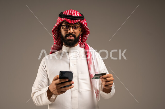  استخدام الأجهزة والتقنيات الحديثة, إمكانية تسديد الفواتير بالبطاقات الإلكترونية, سهولة الشراء والدفع عن طريق بطاقة الإئتمان البنكية، بورتريه مقرب لرجل عربي خليجي سعودي مبتسم يرتدي الثوب التقليدي والشماغ يمسك بيده بطاقة الصراف ويعمل على الهاتف المحمول, خلفية رمادية