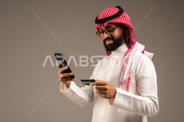  استخدام الأجهزة والتقنيات الحديثة, إمكانية تسديد الفواتير بالبطاقات الإلكترونية, سهولة الشراء والدفع عن طريق بطاقة الإئتمان البنكية، بورتريه مقرب لرجل عربي خليجي سعودي مبتسم يرتدي الثوب التقليدي والشماغ يمسك بيده بطاقة الصراف ويعمل على الهاتف المحمول, خلفية رمادية