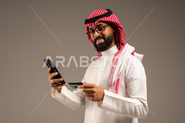  استخدام الأجهزة والتقنيات الحديثة, إمكانية تسديد الفواتير بالبطاقات الإلكترونية, سهولة الشراء والدفع عن طريق بطاقة الإئتمان البنكية، بورتريه مقرب لرجل عربي خليجي سعودي مبتسم يرتدي الثوب التقليدي والشماغ يمسك بيده بطاقة الصراف ويعمل على الهاتف المحمول, خلفية رمادية