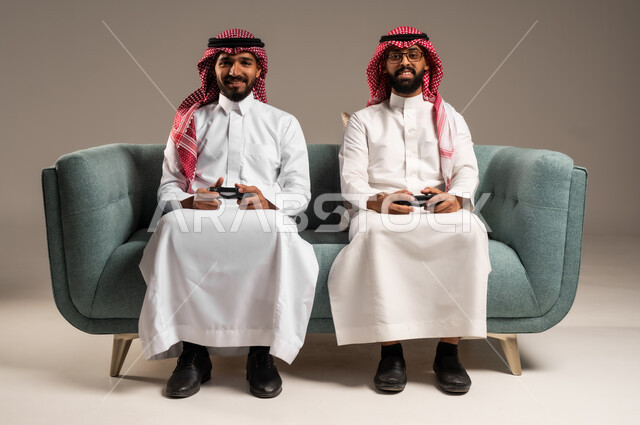 المشاركة والمنافسة بالألعاب التقنية الحديثة والمتطورة,  بورتريه لرجلين عربيين خليجيين سعوديين يرتديان الثوب التقليدي والشماغ يمسكان عصا التحكم, العالم الرقمي الترفيهي, المتعة والتسلية بالفيديو جيمز برفقة الأصدقاء