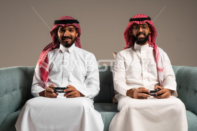 المشاركة والمنافسة بالألعاب التقنية الحديثة والمتطورة,  بورتريه لرجلين عربيين خليجيين سعوديين يرتديان الثوب التقليدي والشماغ يمسكان عصا التحكم, العالم الرقمي الترفيهي, المتعة والتسلية بالفيديو جيمز برفقة الأصدقاء