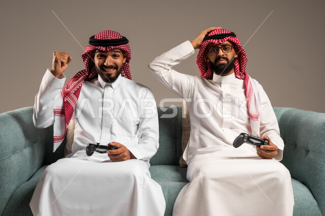 المشاركة والمنافسة بالألعاب التقنية الحديثة والمتطورة,  بورتريه لرجلين عربيين خليجيين سعوديين يرتديان الثوب التقليدي والشماغ يمسكان عصا التحكم, العالم الرقمي الترفيهي, المتعة والتسلية بالفيديو جيمز برفقة الأصدقاء