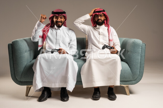 المشاركة والمنافسة بالألعاب التقنية الحديثة والمتطورة,  بورتريه لرجلين عربيين خليجيين سعوديين يرتديان الثوب التقليدي والشماغ يمسكان عصا التحكم, العالم الرقمي الترفيهي, المتعة والتسلية بالفيديو جيمز برفقة الأصدقاء