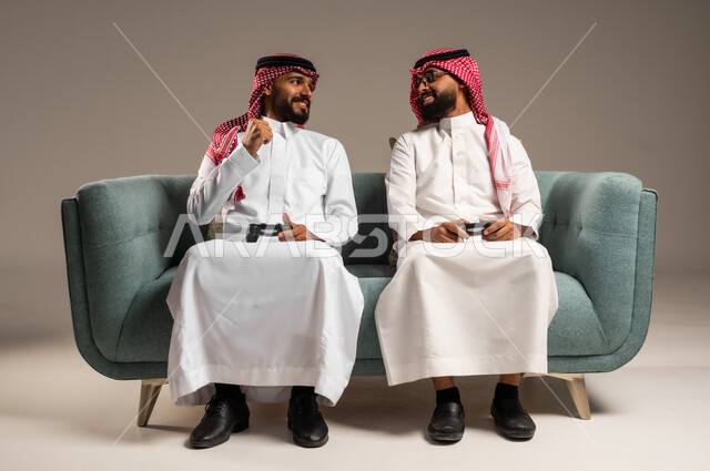المشاركة والمنافسة بالألعاب التقنية الحديثة والمتطورة,  بورتريه لرجلين عربيين خليجيين سعوديين يرتديان الثوب التقليدي والشماغ يمسكان عصا التحكم, العالم الرقمي الترفيهي, المتعة والتسلية بالفيديو جيمز برفقة الأصدقاء
