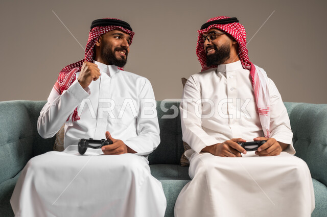 المشاركة والمنافسة بالألعاب التقنية الحديثة والمتطورة,  بورتريه لرجلين عربيين خليجيين سعوديين يرتديان الثوب التقليدي والشماغ يمسكان عصا التحكم, العالم الرقمي الترفيهي, المتعة والتسلية بالفيديو جيمز برفقة الأصدقاء