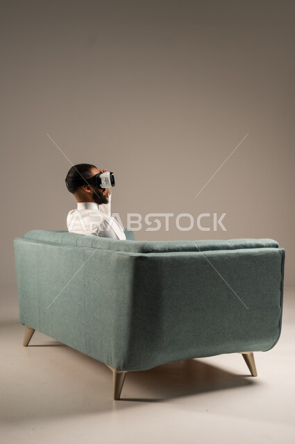 قضاء أوقات ممتعة ومسلية بالألعاب الافتراضية، الاستمتاع بمشاهدة أفلام الواقع الافتراضي، بورتريه من الخلف لرجل عربي خليجي سعودي يرتدي الثوب التقليدي يجلس على كرسي مريح ويرتدي نظارة الواقع الافتراضي ثلاثية الابعاد، خلفية رمادية