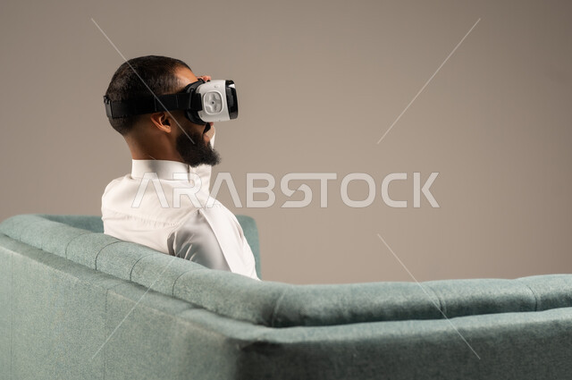 Having fun and entertaining times with virtual games, enjoying watching virtual reality movies, portrait from the back of a Saudi Arabian Gulf man wearing traditional thobe sitting on a comfortable chair and wearing 3D virtual reality glasses, gray background