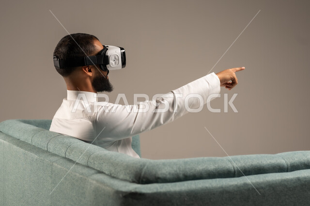 قضاء أوقات ممتعة ومسلية بالألعاب الافتراضية، الاستمتاع بمشاهدة أفلام الواقع الافتراضي، بورتريه من الخلف لرجل عربي خليجي سعودي يرتدي الثوب التقليدي يجلس على كرسي مريح ويرتدي نظارة الواقع الافتراضي ثلاثية الابعاد، خلفية رمادية